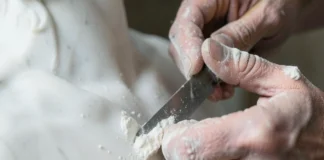 Inquadratura ravvicinata di mani esperte che usano uno scalpello per modellare una statua di marmo sotto una luce soffusa.