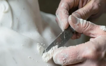 Inquadratura ravvicinata di mani esperte che usano uno scalpello per modellare una statua di marmo sotto una luce soffusa.