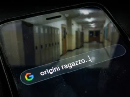 Smartphone in un ambiente oscuro con una ricerca Google sulle origini interrotta e un riflesso di un corridoio scolastico vuoto e sfocato.