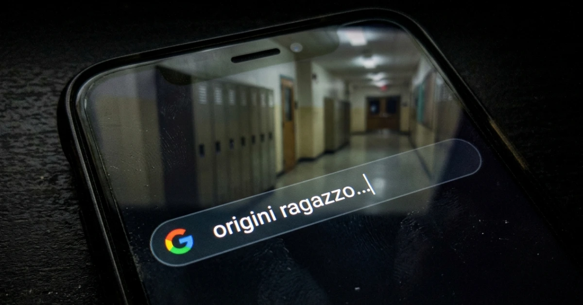 Smartphone in un ambiente oscuro con una ricerca Google sulle origini interrotta e un riflesso di un corridoio scolastico vuoto e sfocato.