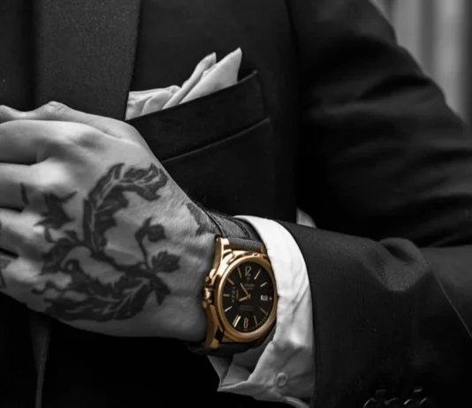 Primo piano cinematografico di una mano tatuata che stringe il bavero di una giacca sartoriale di alta qualità, con un orologio al polso, su uno sfondo di un vicolo urbano sfocato con graffiti.