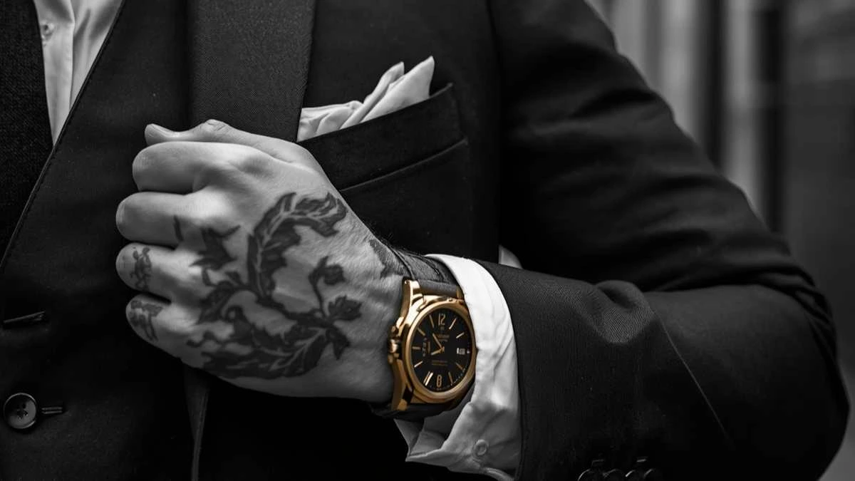 Primo piano cinematografico di una mano tatuata che stringe il bavero di una giacca sartoriale di alta qualità, con un orologio al polso, su uno sfondo di un vicolo urbano sfocato con graffiti.